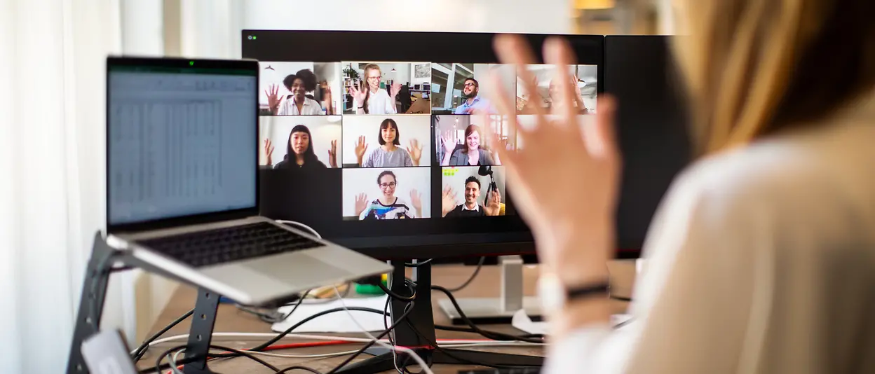 A woman sits in front of her computer and waves to her colleagues, whom she sees on the computer screen during an online meeting. 