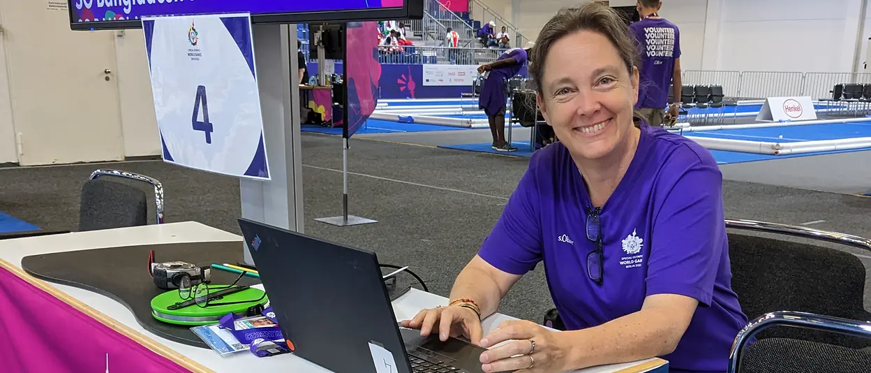 Kirsten sits on a table with a laptop placed in front of her while behind her is a big screen showing information regarding the current boccia match.