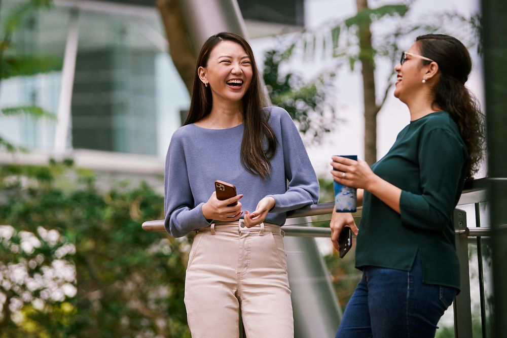 Two women standing in the green outdoor space of a modern office building laughing together.