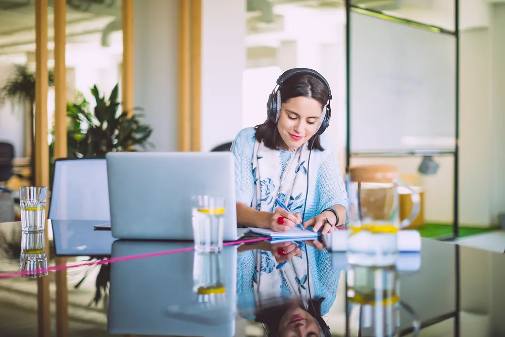 A young woman sitting at a table with her laptop is writing on a notepad.