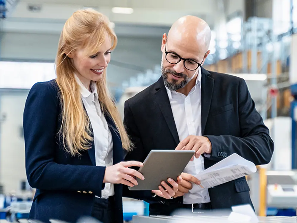 Two business people stand in a manufacturing site and look at a document