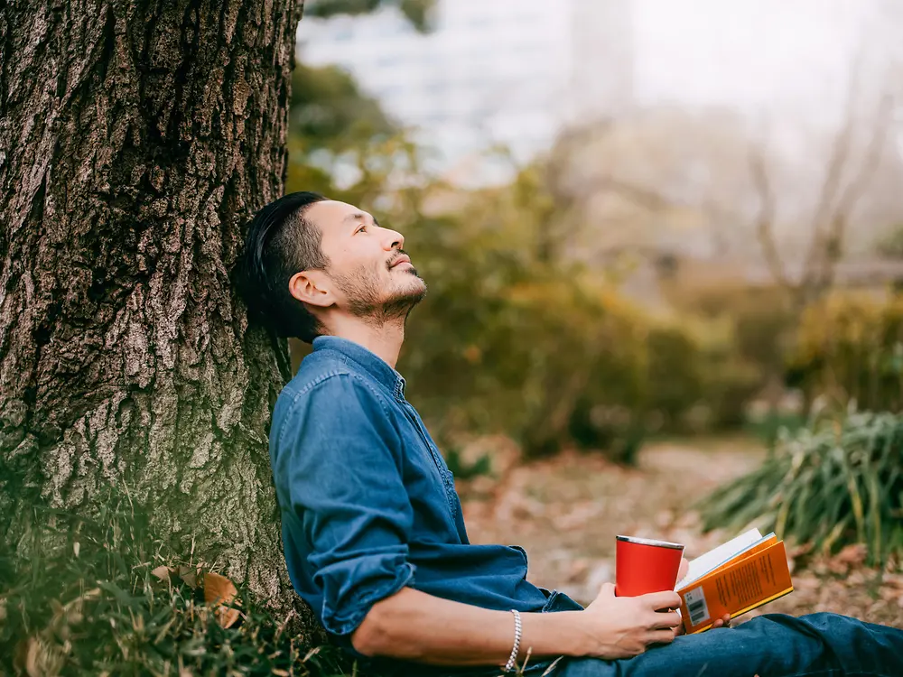 A man with a book leaning against a tree