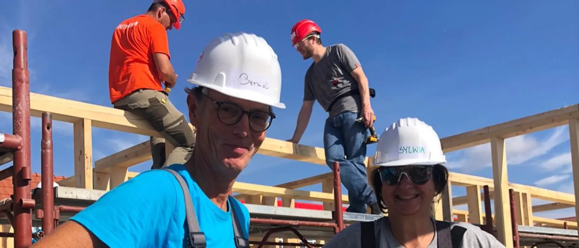 Two volunteers with helmets on their heads smile at the camera and stand in front of a scaffold on which two other helpers are standing.