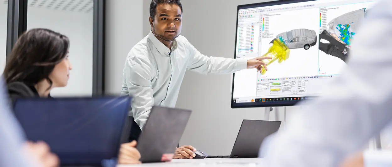 In a meeting with several people in front of their computers, one person in a white shirt is standing up and pointing at 3D models of car on a big screen.