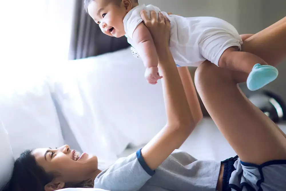 A smiling young woman lays on a white sofa. She is playing with a toddler she is holding in her hands.