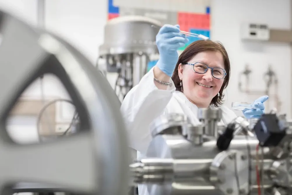 A smiling, female lab technician is wearing a white coat, safety glasses and blue latex gloves. She is holding a small object with some pliers while working in front of a XPS machine