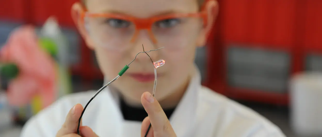 A boy in a lab coat and safety glasses holding a conductivity tester