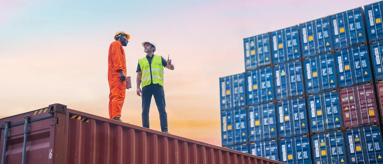 Two men stand on a container at a commercial port and talk to each other.