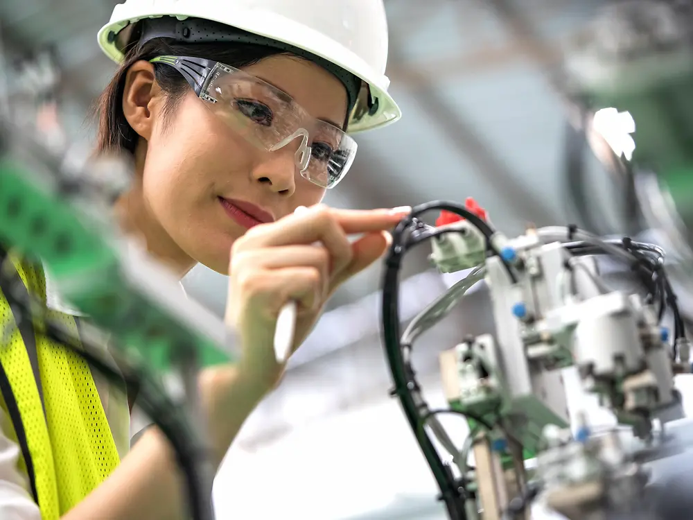 Woman wearing a hard hat and safety glasses inspects industrial machinery