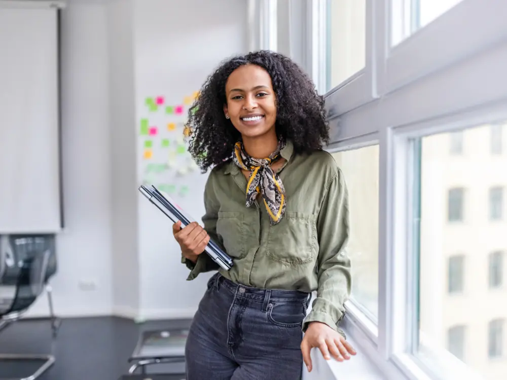 A woman smiling while standing by the window and holding a laptop in one hand.