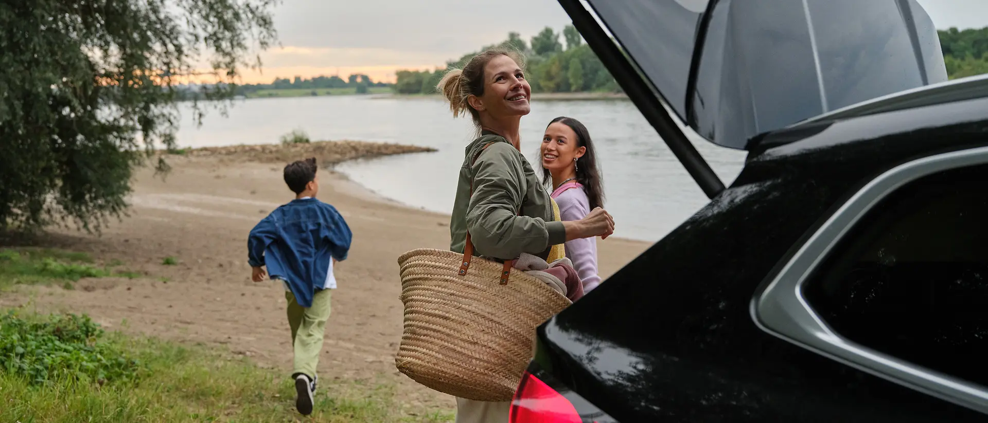 A mother is just about to close the trunk of her car while her daughter watches and her son is already running towards the water on the beach.