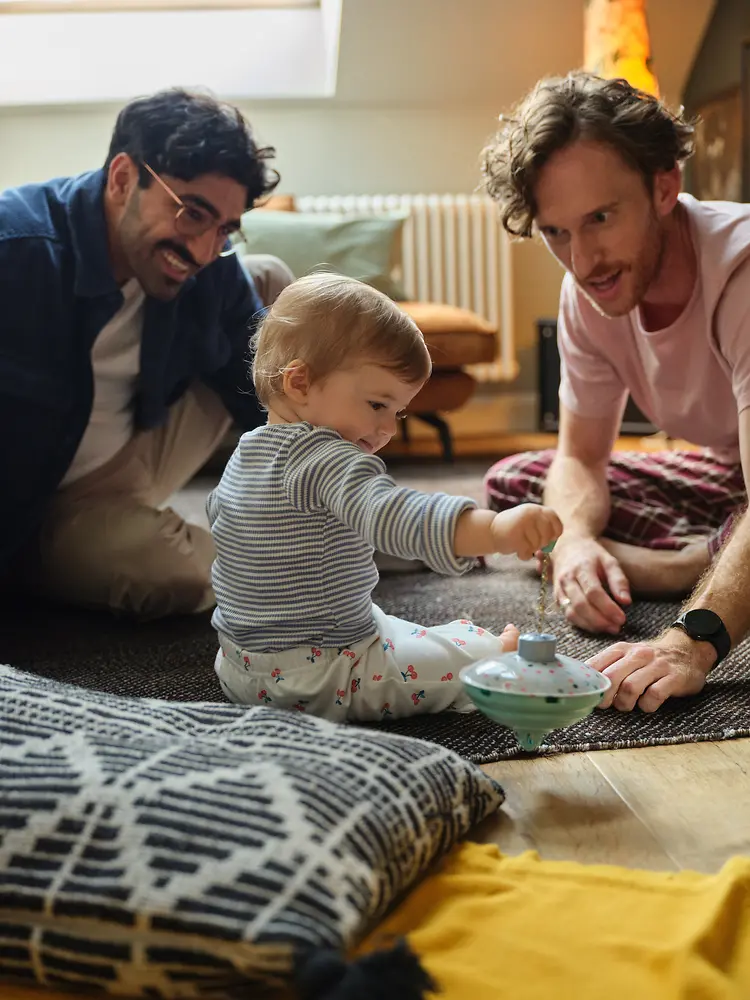 Two male parents are sitting on the floor with their baby daughter, who is focused on playing with a spinning top. 