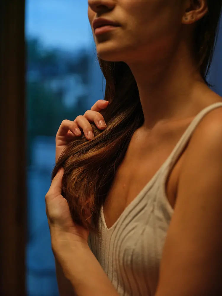 A young woman with long, dark hair is shown from the side as she gently runs her hands through her hair, working in a hair care product.