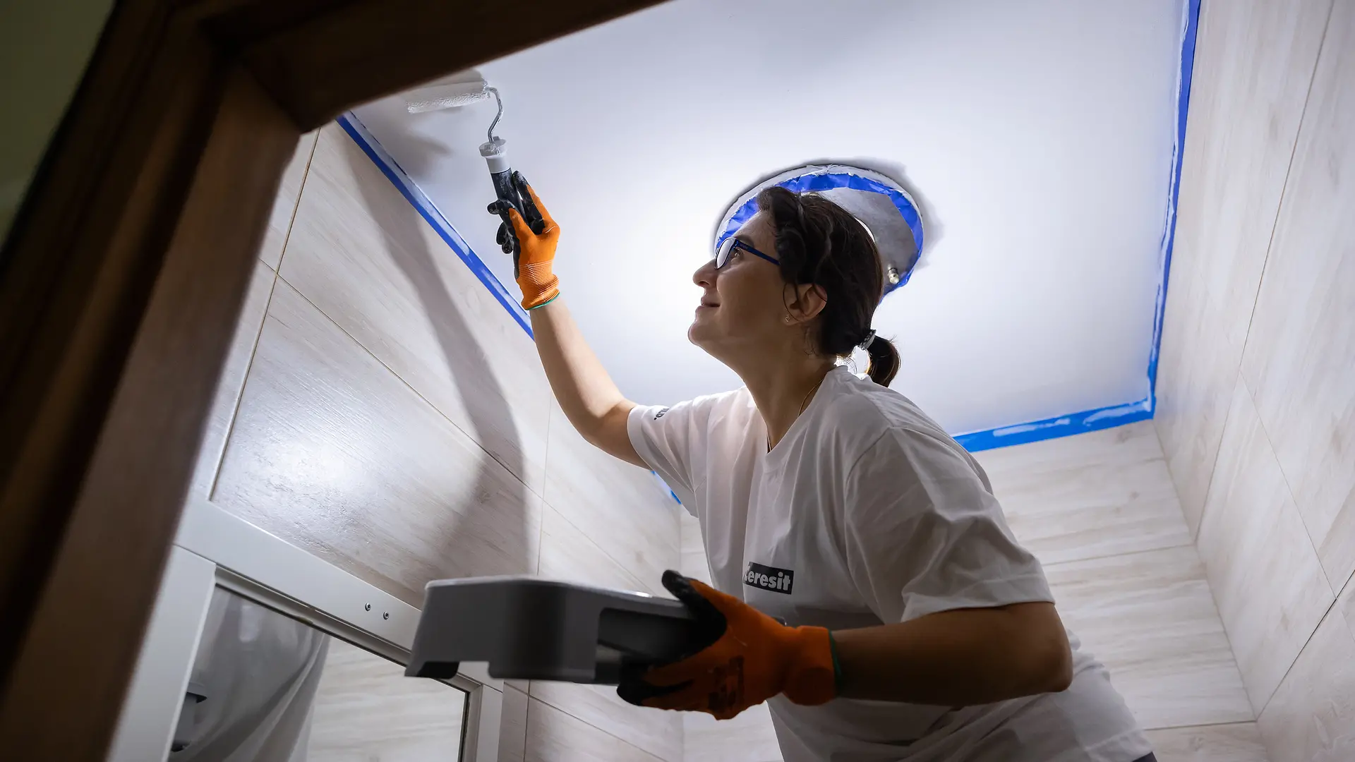 A Henkel employee paints the ceiling of a small room with white paint.