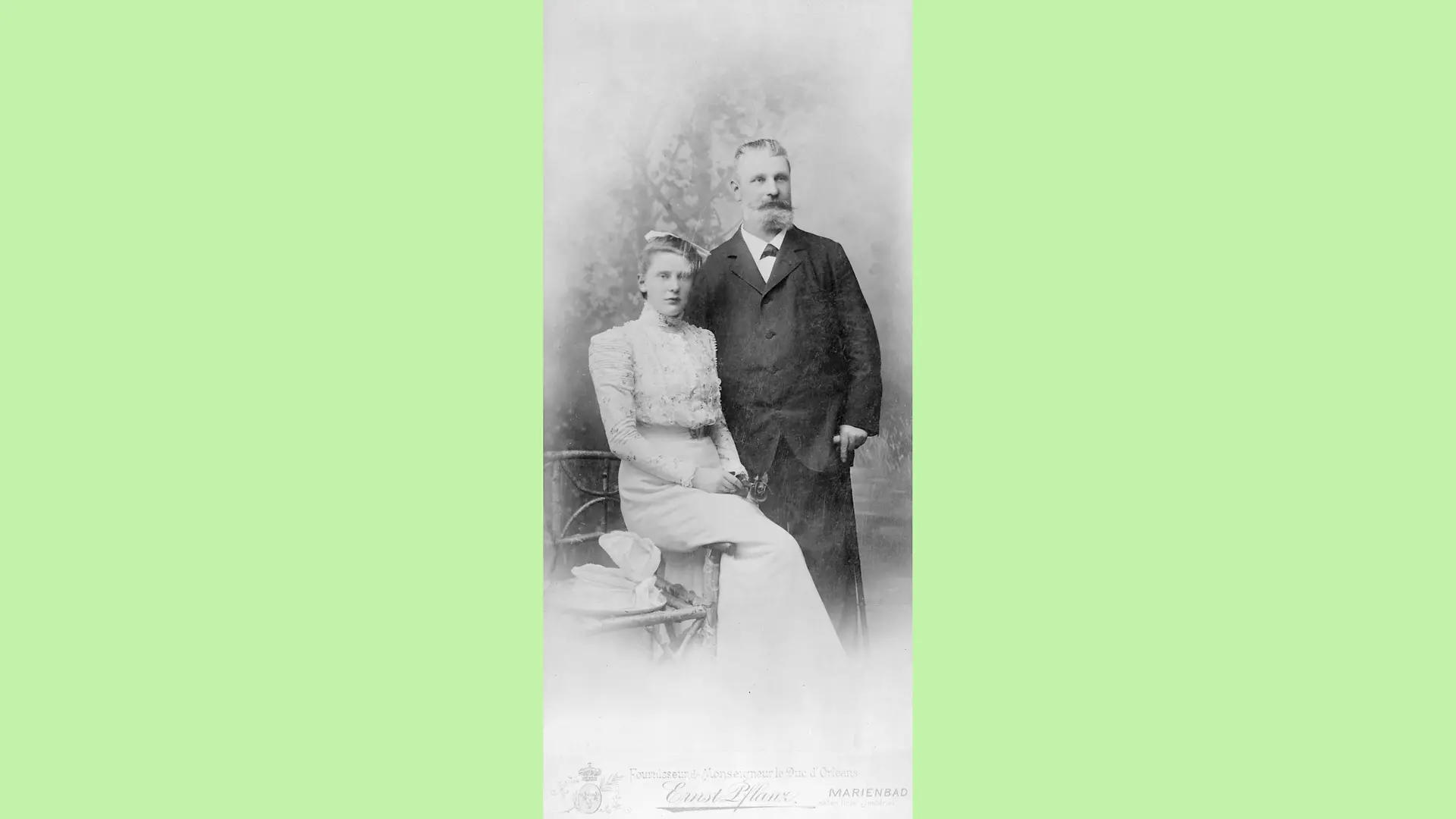 Emmy Lüps in her wedding dress, sitting sideways on the back of a chair. Her father Fritz Henkel is standing next to her, gazing into the distance and holding a cane in his left hand.