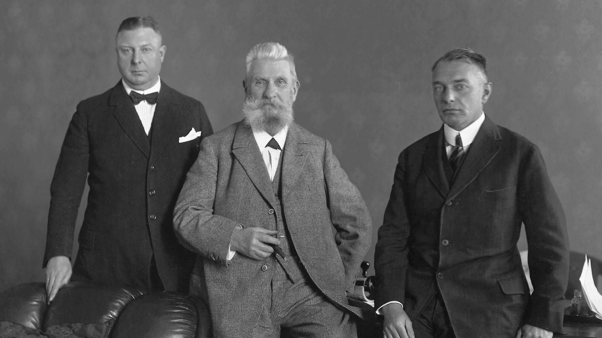 Black and white photograph of Fritz Henkel and his sons Fritz Jr. and Hugo. Fritz Henkel senior is placed half-sitting/reclining in the center. Fritz Jr. is standing at his right side, his legs are covered by a leather sofa. Hugo is leaning against a table on his father's left.