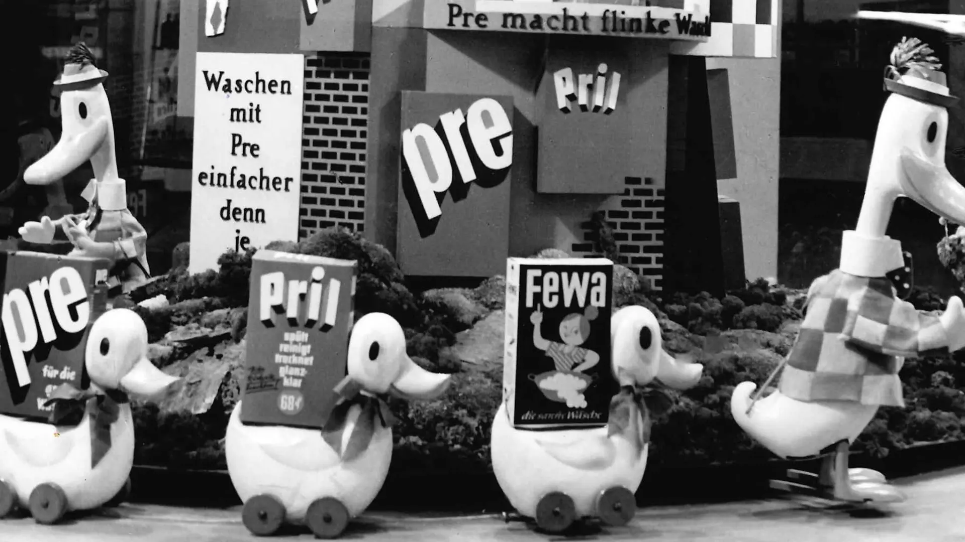 Advertising campaign for Pril and other Henkel products with oversized duck figures. The figures stand in a semicircle and carry various oversized packages of Henkel products on their backs. Behind the figures are many advertising posters for the corresponding products.