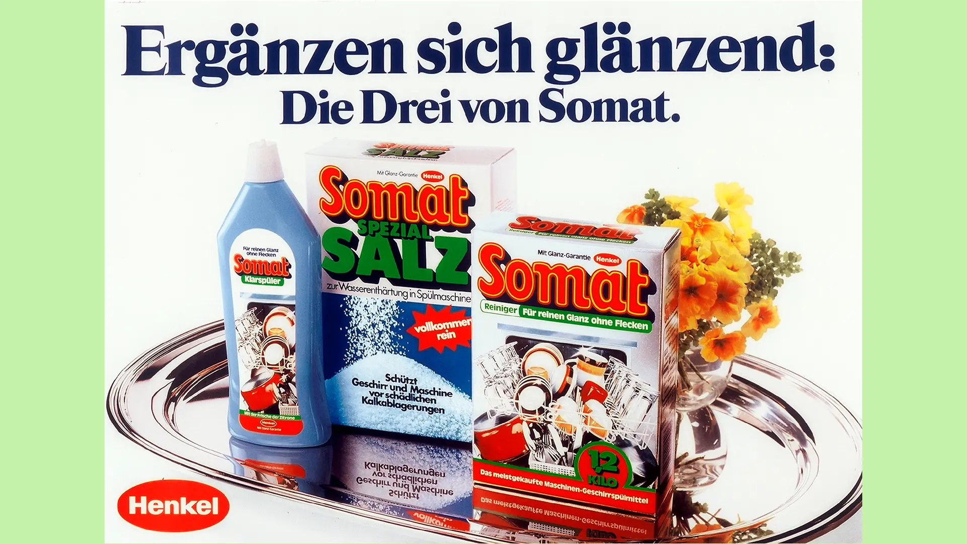 Somat rinse aid, special salt and detergent slightly offset and placed next to each other on a silver tray. In the background, half hidden by the Somat detergent, is a small orange bouquet of flowers in a bulbous glass vase. The slogan “Complement each other brilliantly: The three from Somat” can be read above it.n blouse smiles as she empties her clean dishwasher. In front of the waiter in the bottom right-hand corner, the packaging of Somat detergent and rinse aid is also depicted.