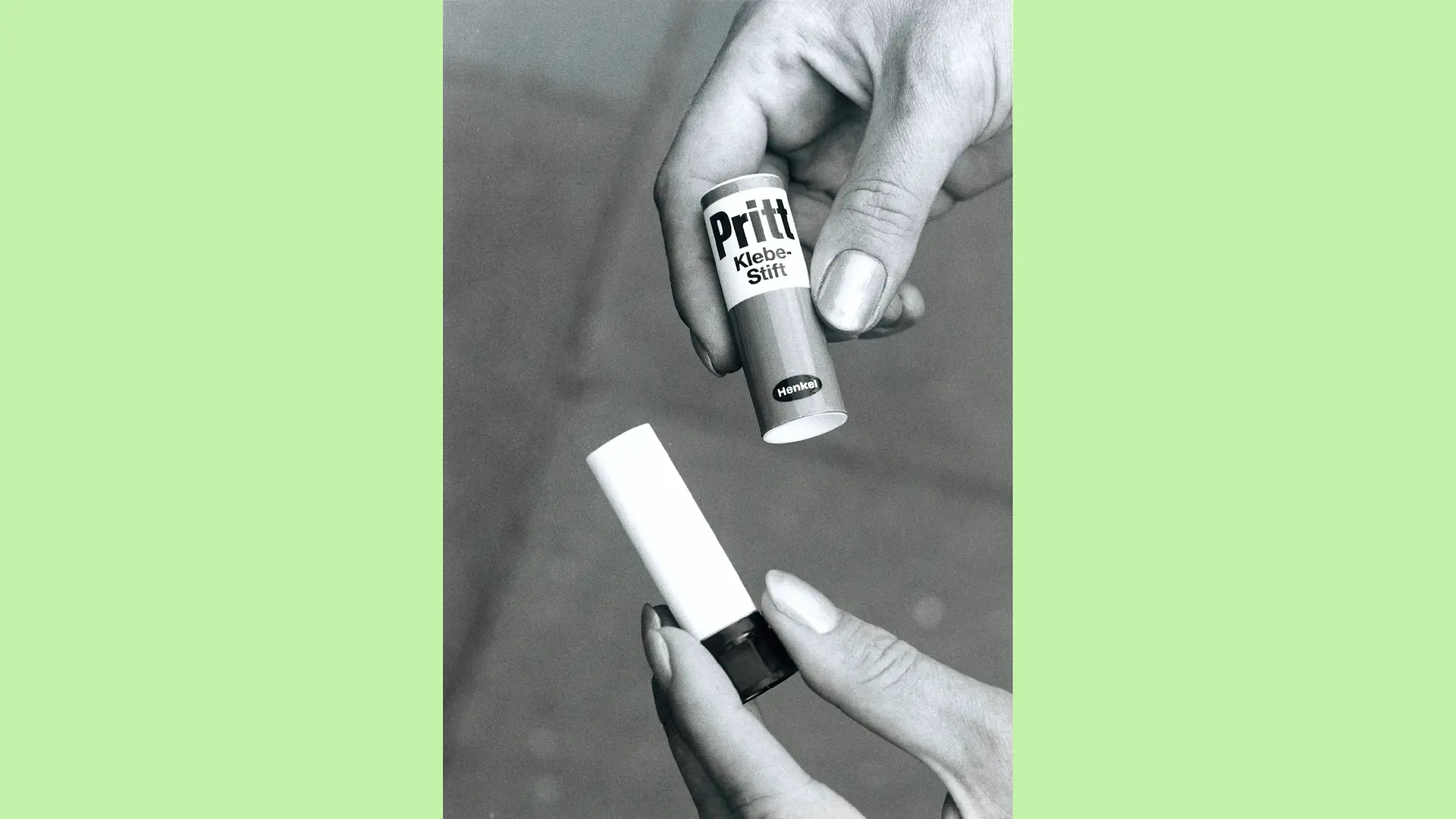 Black and white photo of hands holding an open Pritt glue stick. The right hand holds the lid while the left hand holds the part with the twist mechanism.