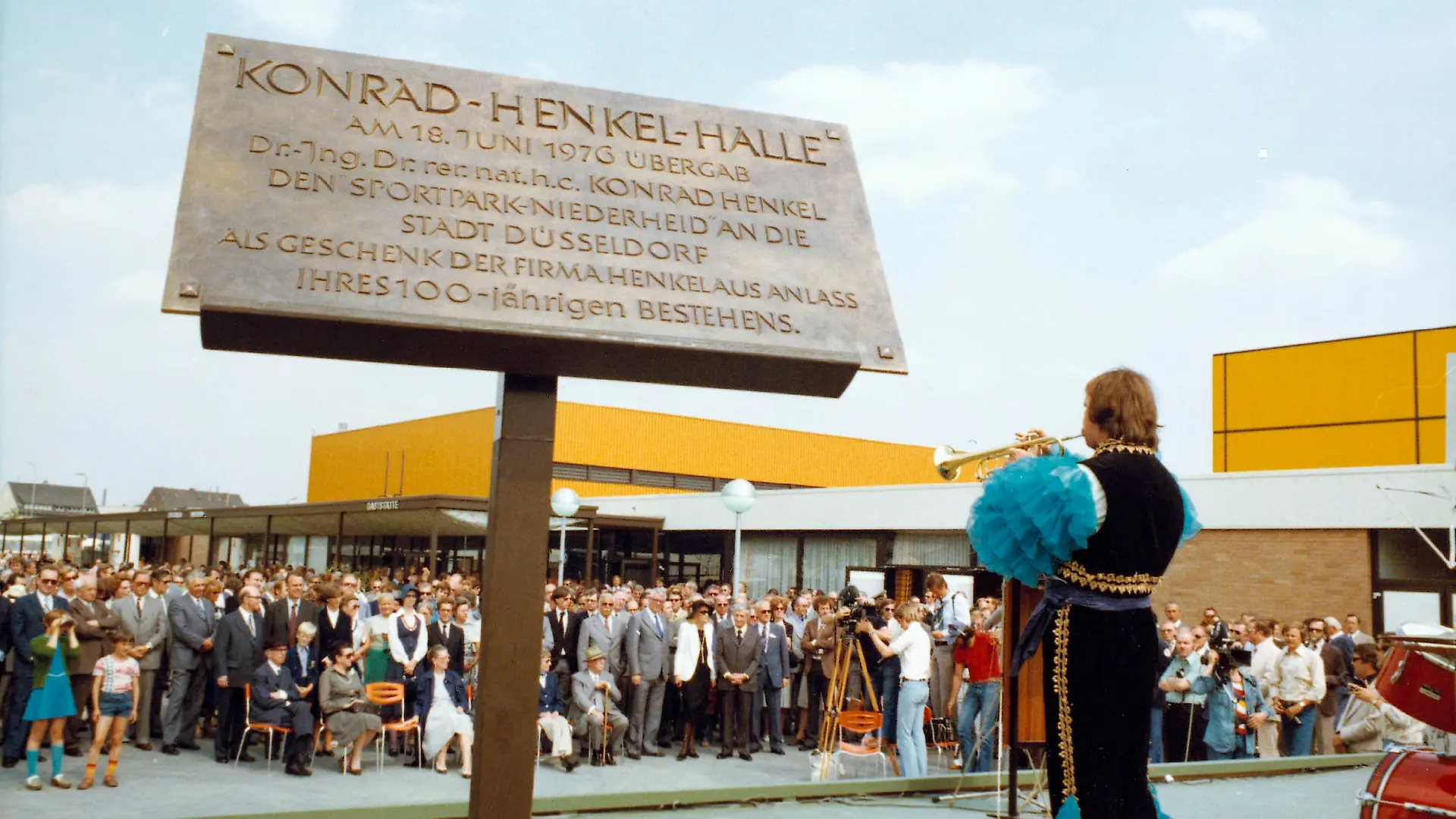 Photo from the opening ceremony of Sportpark Niederheid. In the foreground, a commemorative plaque in honor of the Henkel company for the donation of the sports park to the city of Düsseldorf on the occasion of the company's 100th anniversary. A man, his back turned to the camera and wearing a striking costume with blue ruffles on his arms and legs, plays a trumpet in front of an assembled audience.
