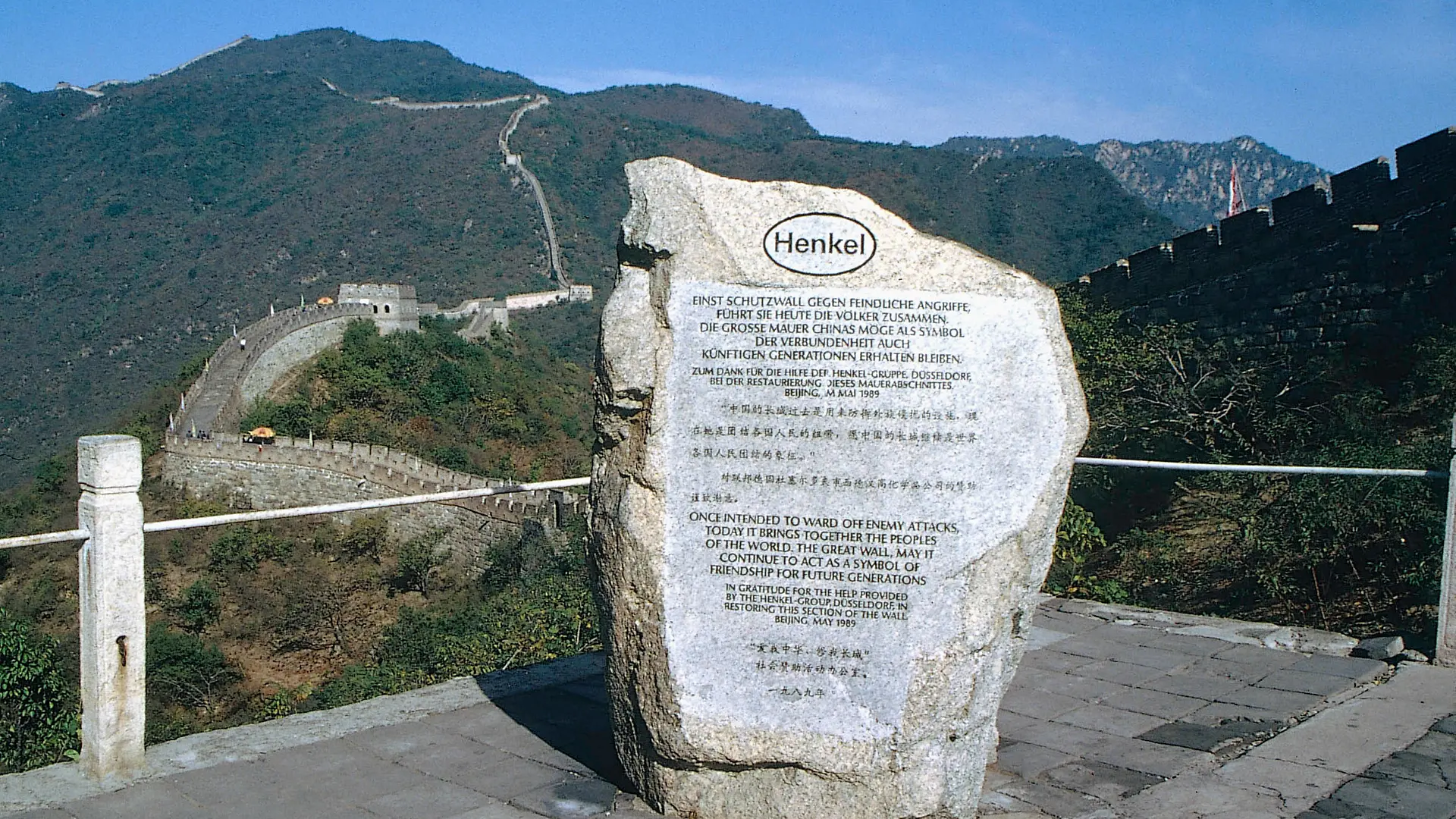 A memorial stone commemorating Henkel's support for the restoration of a section of the Great Wall of China. In the background, the Great Wall of China winds its way through a forested mountainous section.