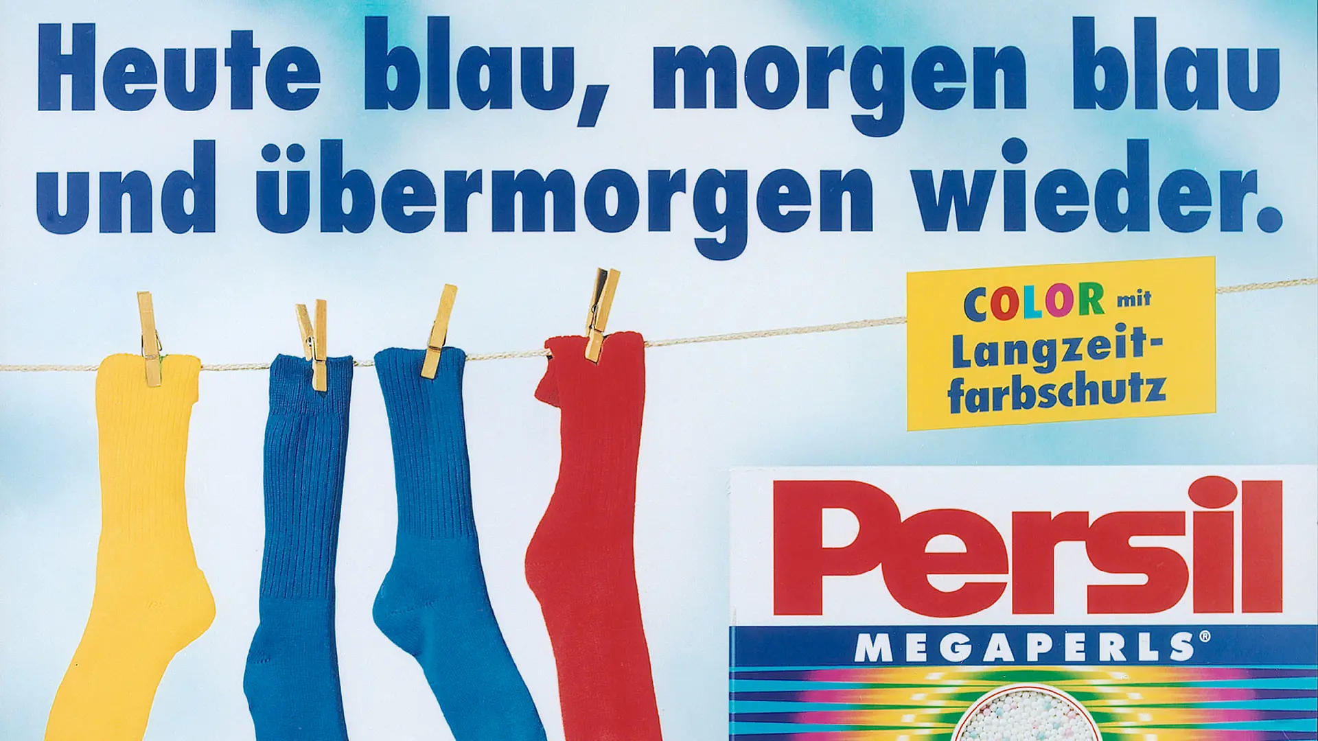 An advertisement for Persil Color. A washing line with four colorful socks, including two blue ones, hangs in the background of the picture. A pack of Persil Color is shown in the right-hand corner and above it is the phrase “Color with long-term color protection”. Above the advertisement is the sentence “Blue today, blue tomorrow and again the day after tomorrow”.