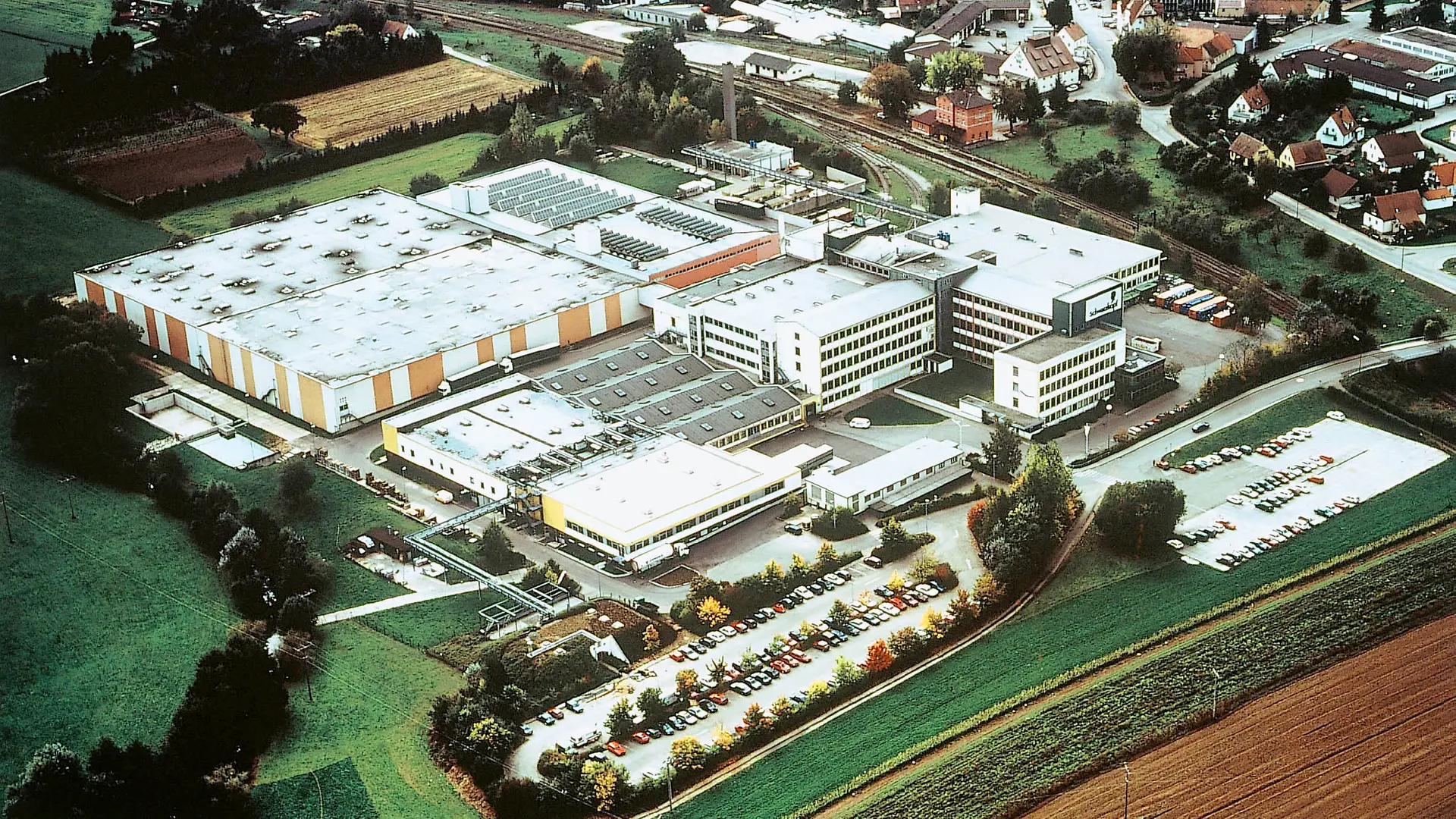 An aerial view of the Schwarzkopf plant in Wassertrüdingen in 1995, showing several production buildings and halls, as well as a large parking lot full of cars.
