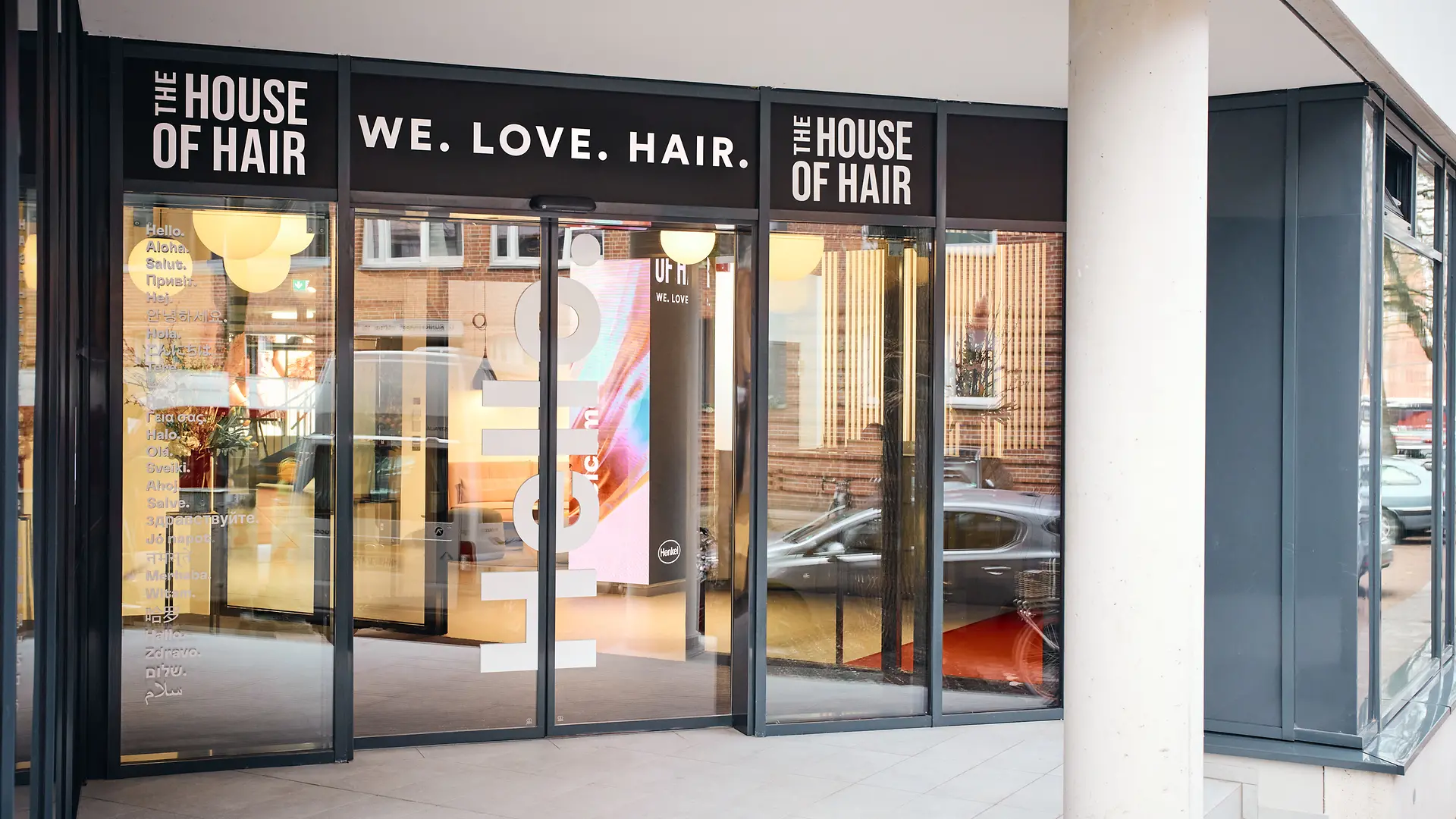 Glazed entrance area. Above the door is the slogan “We. Love. Hair.” To the right and left of it is the inscription “The House of Hair.”